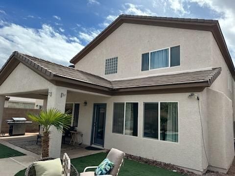 Beige two-story house with brown roof and trim, several windows, and a covered patio with a grill.