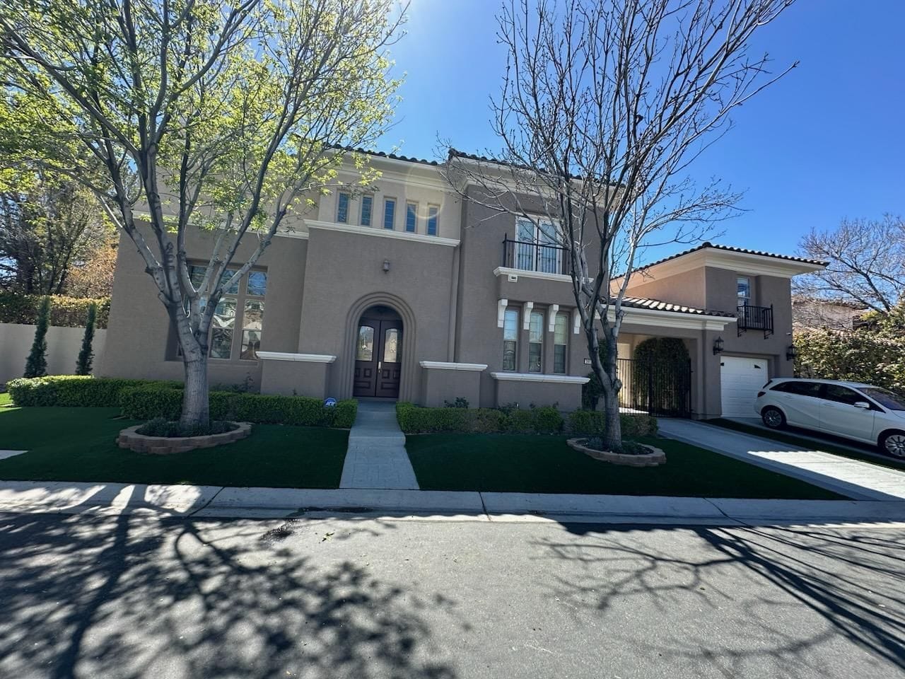 Two-story stucco house with arched doorway and garage. Green lawn and trees in front on a sunny day.