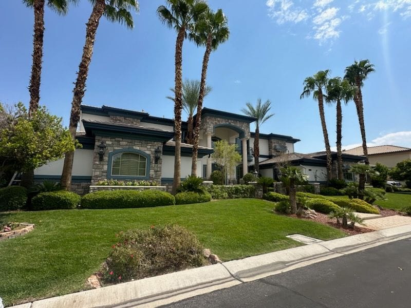 Large home with stone facade, palm trees, and manicured lawn under a blue sky.