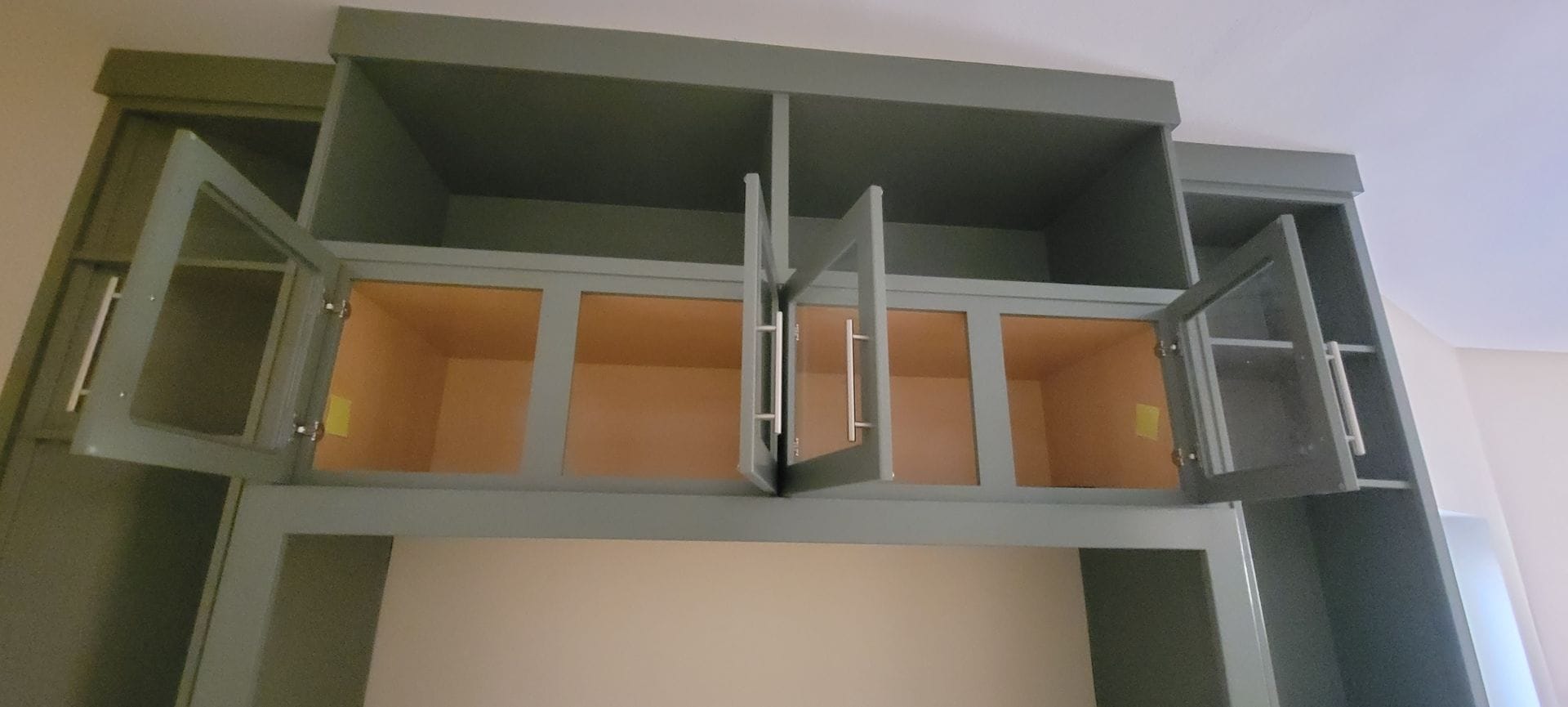 Open, empty kitchen cabinets with glass doors. Cabinets are gray. Interior walls are beige.